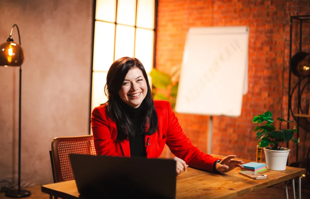 Lachende Frau im roten Blazer am Holztisch vor Backsteinwand und Flipchart