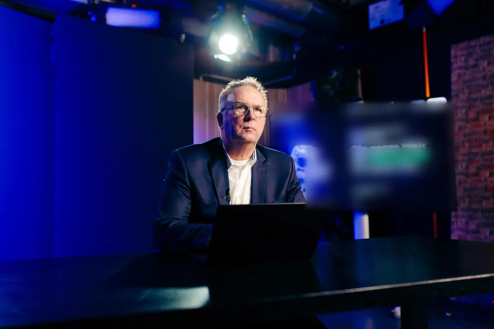 Dunkelblaues Set mit Tisch und Monitor im Hintergrund, Mann im Business-Look